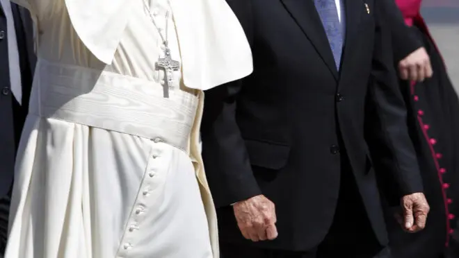 Papa Francisco ao lado de um homem vestido de terno preto com gravata azul. Ele acena para câmera e veste roupas brancas. Suas vestes deixam claro a resposta para a pergunta de quanto ganha um papa, devido sua humildade.