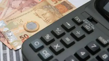 Calculadora ao lado de notas de cinquenta e dez reais, além de uma moeda de um real.